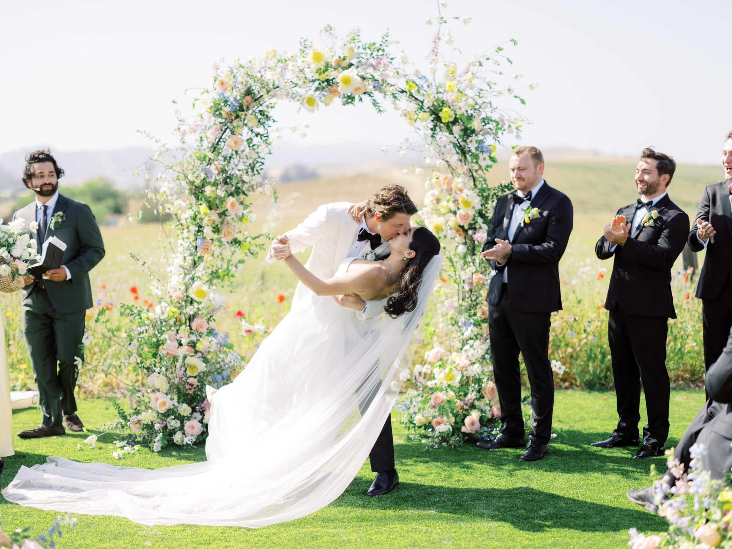 A newlywed couple shares a kiss in front of a floral arch, surrounded by their wedding party dressed in black suits, on a grassy outdoor setting.