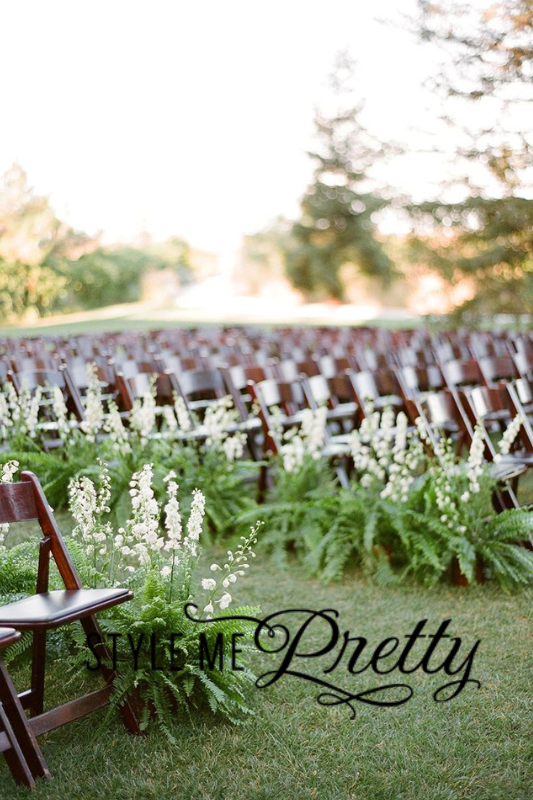 An outdoor wedding ceremony setup with rows of wooden chairs surrounded by lush green plants and white flowers.