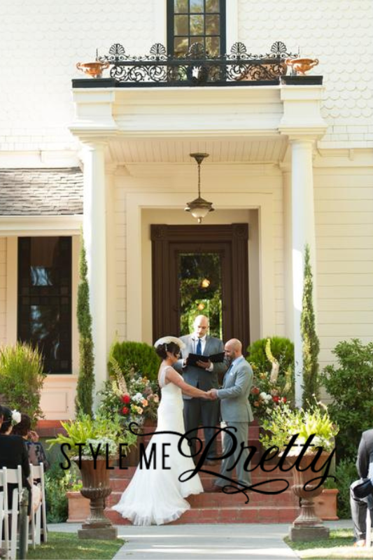 A bride and groom exchange vows outdoors in front of a building with an officiant standing behind them. The bride wears a white dress, and the groom wears a gray suit.