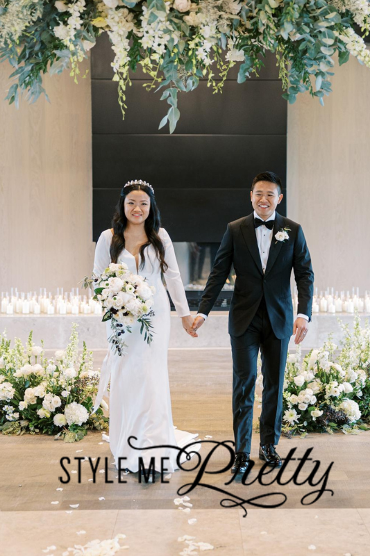 A bride in a white dress and groom in a black tuxedo hold hands and smile while walking down an aisle decorated with white flowers and greenery. The text "STYLE ME Pretty" is overlaid at the bottom.