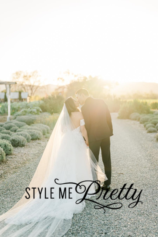 A couple dressed in wedding attire shares a kiss on a gravel path with the sun setting in the background. Overseen by renowned Tahoe wedding planners, the image is overlaid with the text "Style Me Pretty.