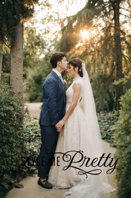 A groom in a blue suit kisses the forehead of his bride in a white gown and veil as they hold hands on a garden path. Sunlight filters through the trees in the background.