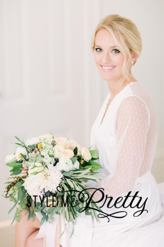 A woman in a white dress, holding a bouquet of flowers, smiles while seated. The image is watermarked with "Style Me Pretty".