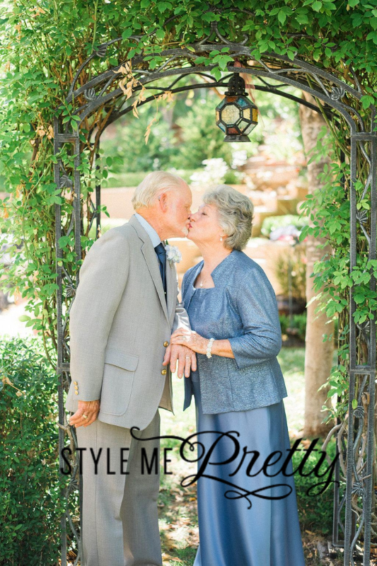 An elderly couple, dressed formally, shares a kiss under a decorative garden arch. The woman wears a blue dress and the man wears a light grey suit. The garden is lush and green. "Style Me Pretty" is written on the image.