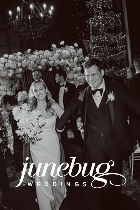 A smiling couple in wedding attire walks amid guests at a wedding celebration, surrounded by floral arrangements and twinkling lights. "junebug WEDDINGS" text is at the bottom of the image.