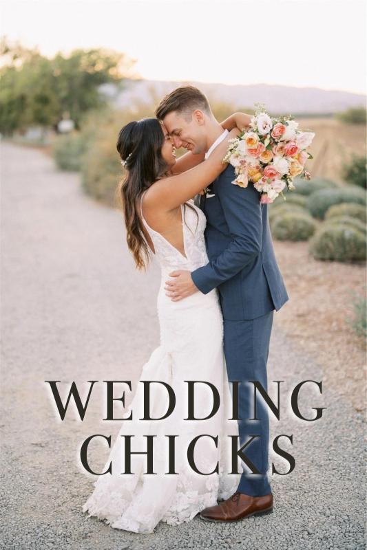 A bride and groom embrace on a gravel path, the groom in a blue suit and the bride in a white dress holding a bouquet of flowers, with "WEDDING CHICKS" text overlaying the image.