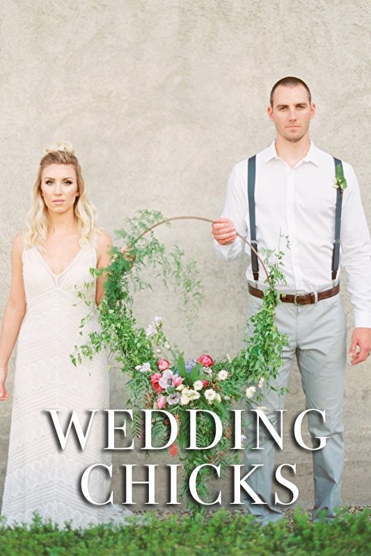 A couple stands side by side. The woman in a white dress and the man in a white shirt with suspenders, holding a green wreath with flowers. Text at the bottom reads "WEDDING CHICKS.