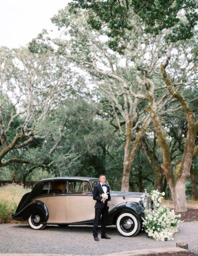 A person in formal attire stands beside a vintage car adorned with white flowers, surrounded by trees.