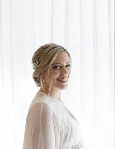 A smiling woman in a bridal gown stands in front of sheer curtains, looking over her shoulder.