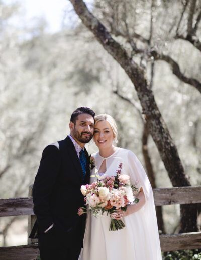 A couple poses for a wedding photo outdoors, with the groom in a suit and the bride in a white dress holding a bouquet of flowers, standing in a wooded area.