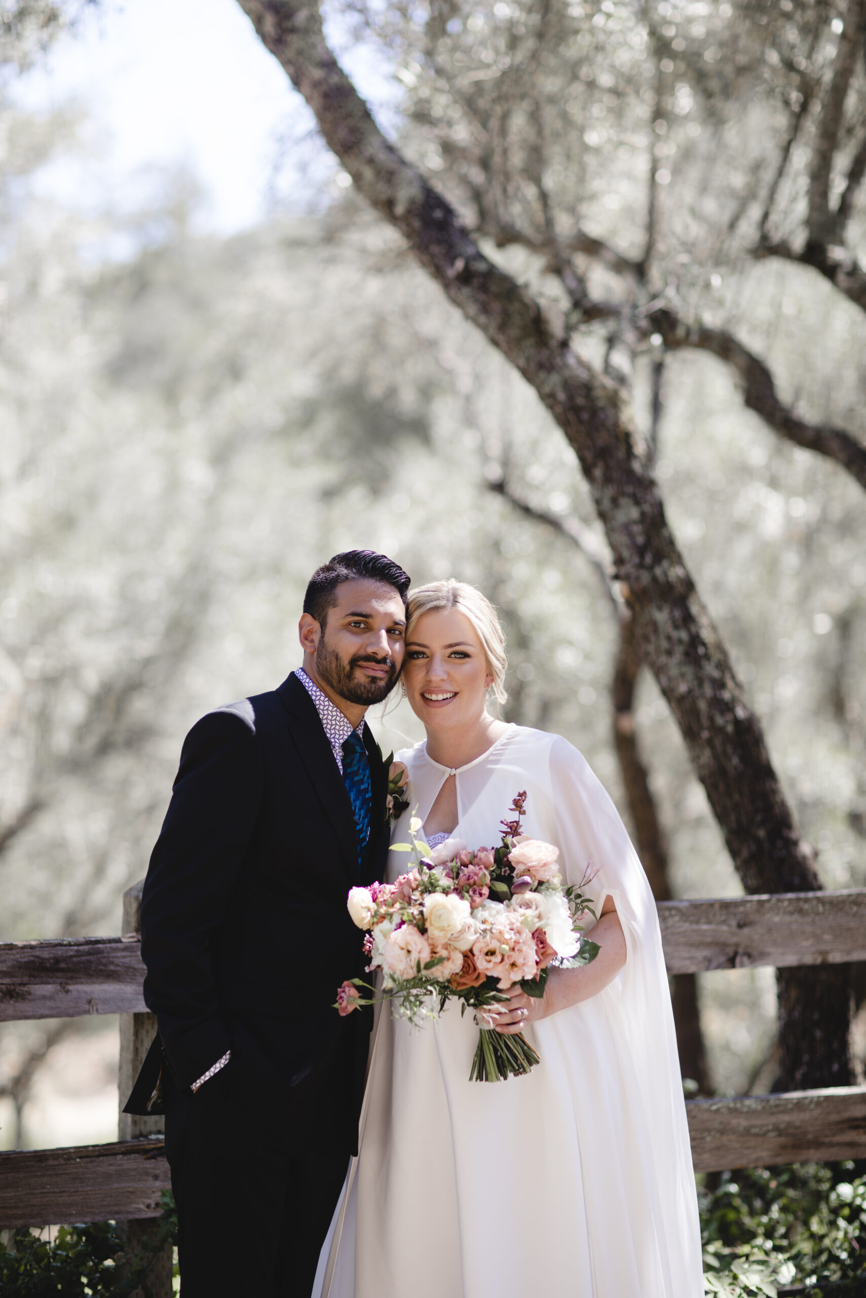 A couple poses for a wedding photo outdoors, with the groom in a suit and the bride in a white dress holding a bouquet of flowers, standing in a wooded area.