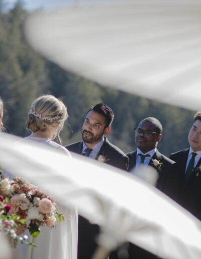 A wedding ceremony with a bride and groom standing before each other, surrounded by guests and groomsmen. Bouquets and white parasols are partially visible in the foreground.