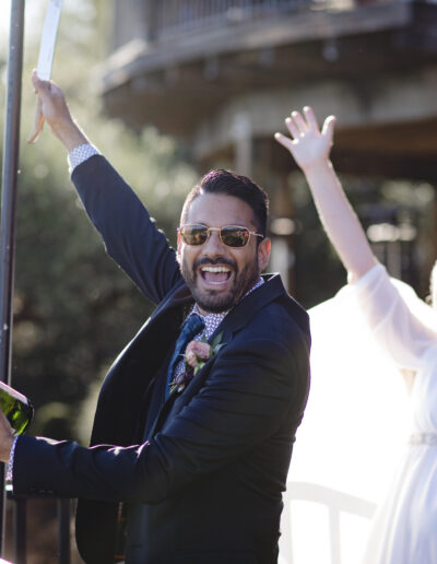 A man in a suit joyfully opens a bottle of champagne while a woman in a white dress raises her arms in celebration outdoors.