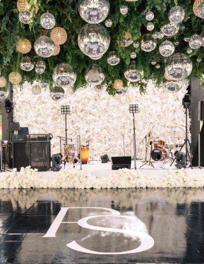 Elegant outdoor stage with white floral backdrop, musical instruments, and reflective spheres hanging from greenery above a glossy black floor with initials "PS.