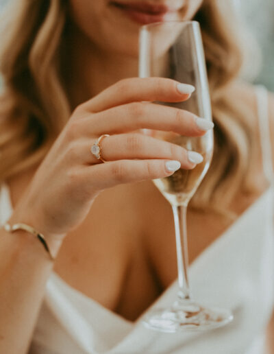 A woman in a white dress holds a glass of champagne, displaying an engagement ring.