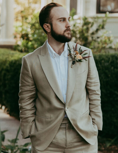 Man in a beige suit with a floral boutonniere stands outdoors, looking to the side.