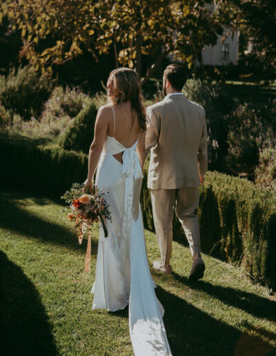 A bride and groom walk through a garden. The bride is holding a bouquet, and wearing a white dress with a large bow at the back. The groom is in a beige suit.