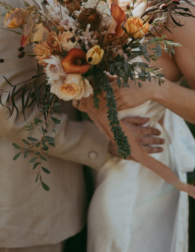 A couple in light-colored attire holding an autumn-themed bouquet with orange and white flowers.