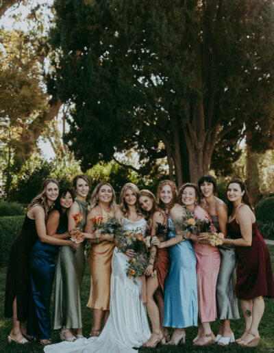 A bride in a white dress stands with a group of bridesmaids in colorful dresses, posing outdoors in front of trees and greenery.