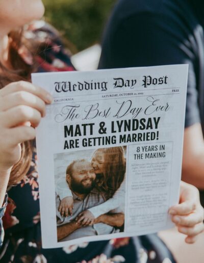 Person holding a newspaper-style wedding announcement for Matt and Lyndsay, titled "The Best Day Ever," with a description of 8 years together.