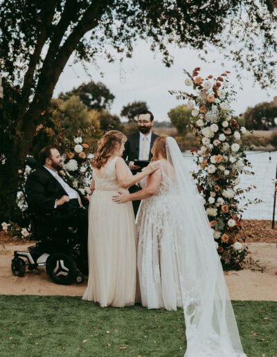 A wedding ceremony by a lake with two brides in white dresses and a person in a suit officiating. One guest, seated in a wheelchair, is part of the event. Floral arch in the background.