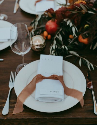 A dining table setting with a white plate, menu, and napkin tied with a brown ribbon. A wine glass is to the left, surrounded by autumn-themed decorations.