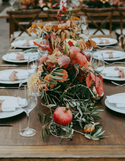 A wooden table set for a meal with white plates, napkins, and glasses. A centerpiece of flowers and foliage runs along the middle, featuring a pomegranate and greenery.