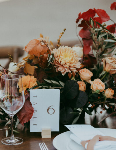 A table setting with a floral centerpiece of autumn-colored flowers, a table number card reading "6," a wine glass, a white plate, and a folded napkin.