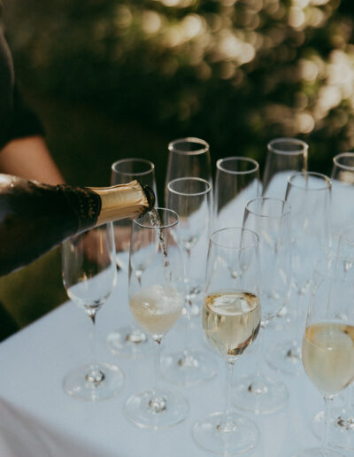 A person pours champagne from a bottle into several glasses on a white tablecloth outdoors.