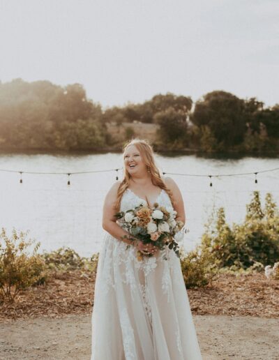 A bride in a white dress stands outdoors, smiling while holding a bouquet. Behind her, a body of water and trees under the sunlight are visible.