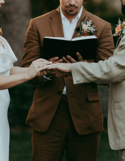 A couple exchanges rings during an outdoor wedding ceremony, with an officiant holding an open book in the background.