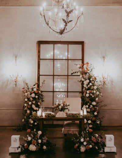 A wedding table is elegantly decorated with floral arrangements, candles, and a chandelier overhead. A backdrop of wooden window panes adds a rustic touch.