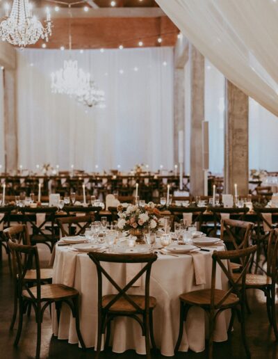 Elegant wedding reception with a round table set for guests, featuring floral centerpieces and lit candles. Chandeliers and draped fabric create a soft, romantic ambiance.