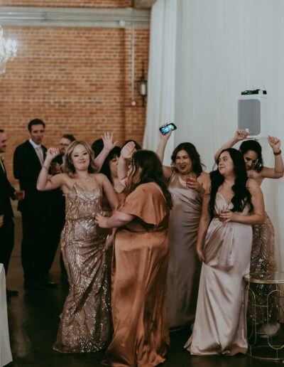 A group of people in formal attire are dancing and smiling at an indoor event with brick walls in the background.