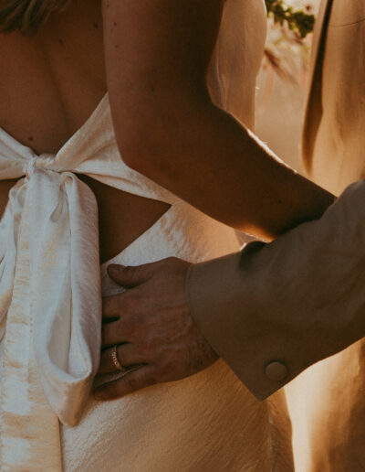 A couple stands closely, with a focus on their hands gently placed on each other's backs. The woman wears a backless white dress; the man's hand is visible with a ring.