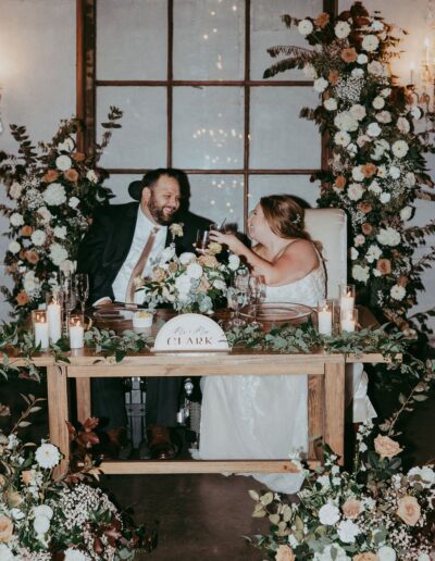 A couple in wedding attire clinks glasses while seated at a table adorned with flowers and candles. A sign reads "Mr. & Mrs. Clark.