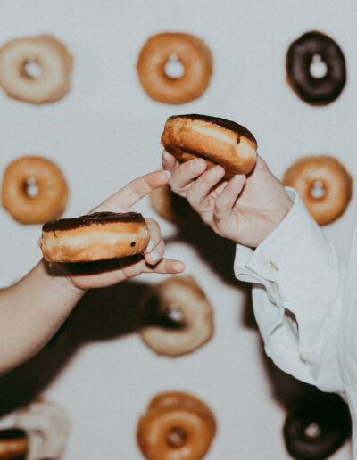 Two people holding chocolate-topped donuts with a wall display of assorted donuts in the background.