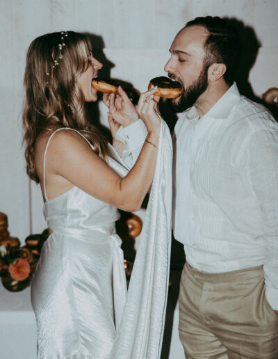 A couple in formal attire feeds each other donuts, standing close in a room with a white wall and floral decor.