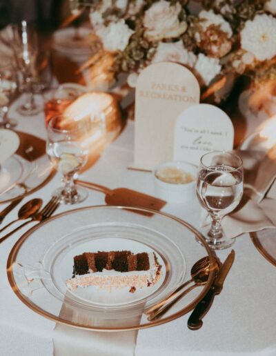 A slice of chocolate cake on a glass plate sits on an elegant table with a floral centerpiece, glassware, utensils, and a "Parks & Recreation" card.