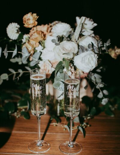 Two champagne flutes engraved with numbers and text are placed in front of a floral bouquet with white and peach roses on a wooden surface.