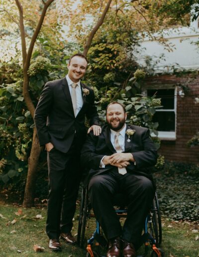 Two men in suits, one sitting in a wheelchair and the other standing beside him, pose for a photo in a garden with trees and greenery.