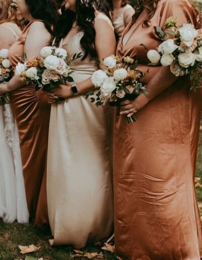 Group of women and a young girl in formal dresses holding bouquets, standing outdoors on grassy ground with fallen leaves.