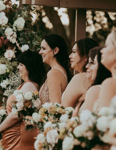 A group of women in formal dresses hold floral bouquets, standing outdoors near a wooden structure adorned with more flowers.