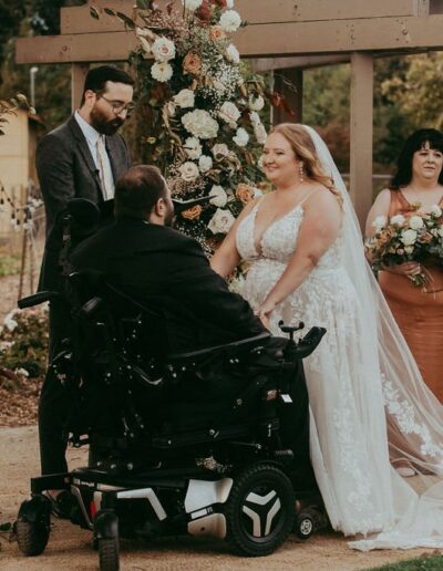 Bride and groom exchange vows under a floral arch, surrounded by bridesmaids holding bouquets. The groom is in a wheelchair.