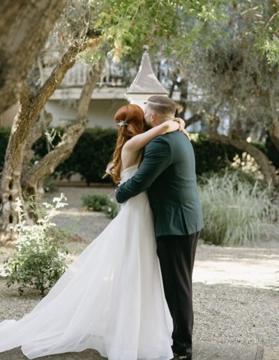 A bride and groom embrace outdoors, surrounded by trees and greenery.