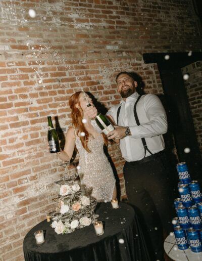 A couple happily sprays champagne in front of a brick wall, standing next to a table with flowers, candles, and a stack of beer cans.