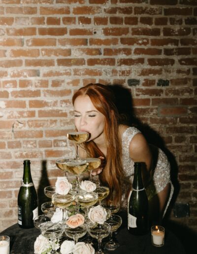 A woman in a dress leans in to drink from a top champagne glass of a pyramid stack. Bottles and candles are on a nearby table against a brick wall.