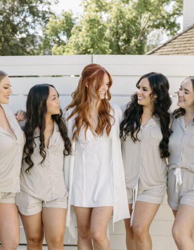 Seven women in matching loungewear stand together outdoors, with one woman in a white robe at the center, smiling and facing each other.