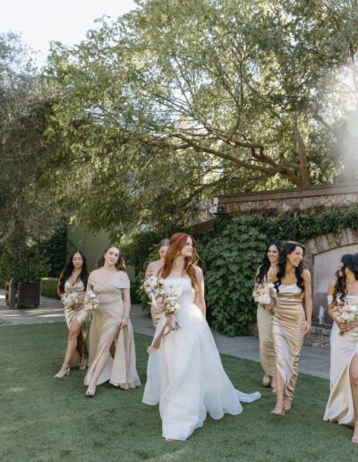 A bride in a white dress walks with bridesmaids in gold dresses on a grassy area, surrounded by trees and greenery.