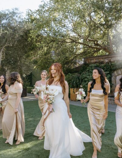 A bride in a white dress walks with bridesmaids in gold dresses on a lawn, surrounded by trees.